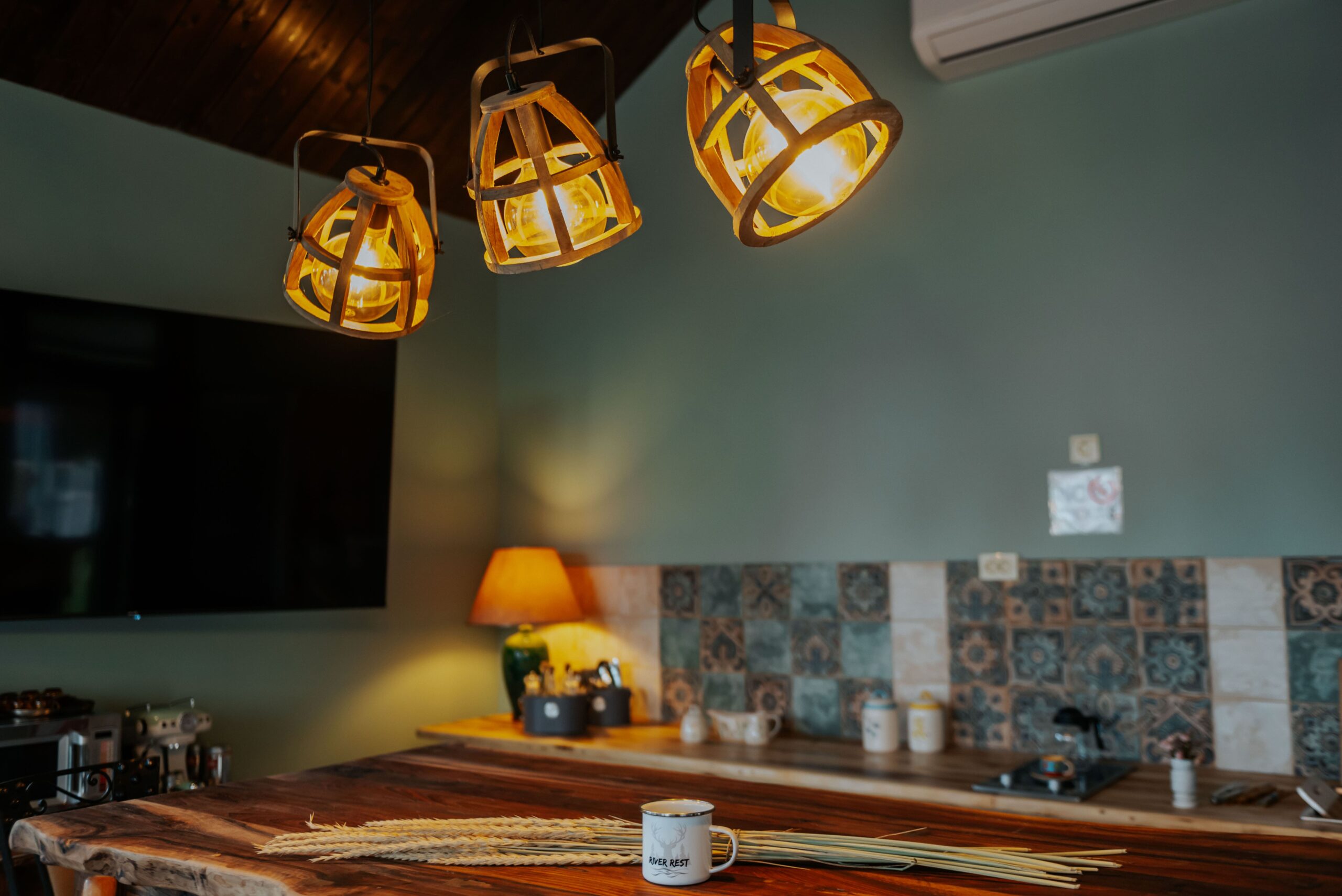 Уютен интериор в River Rest Vetren с дървени лампи, кухненски кът и чаша върху масивна дървена маса.